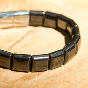 Op de afbeelding: Een armband bestaande uit gladde, vierkante zwarte kralen met een reflecterende, metallic glans. Het sieraad is voorzien van een gepolijste zilverkleurige metalen sluiting die de uiteinden van de kralenstreng verbindt tot een cirkelvormig ontwerp.