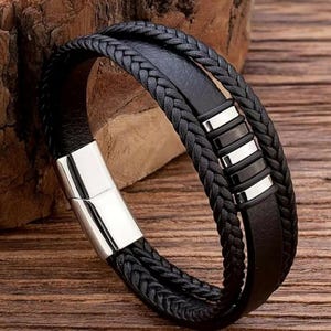 May include: A black leather bracelet with a braided design and a silver metal clasp. The bracelet has a row of silver and black rectangular accents.