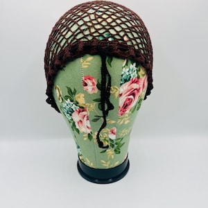May include: A dark brown crocheted hair net with a diamond pattern, displayed on a floral-patterned mannequin head. The net has a scalloped edge and a drawstring closure. The mannequin head is covered in a green fabric with pink and white roses.