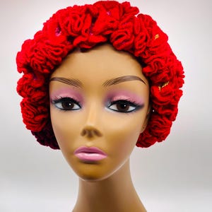 May include: A vibrant red, handmade head wrap with a textured, floral design. The head covering is displayed on a mannequin head with makeup, showcasing the intricate details of the fabric and style. The head wrap is a bold fashion statement.