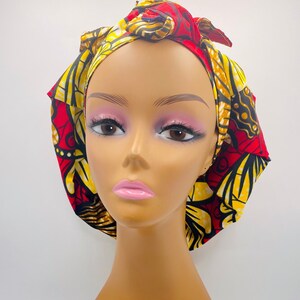 May include: A red and yellow patterned satin hair bonnet with a knot on top. The bonnet is worn on a mannequin head.