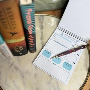 May include: A white book club notes notepad with a spiral binding. The notepad has sections for title, author, rating, meet date, favorite and least favorite parts, most compelling character, interested or frustrated, notes, and was the ending satisfying. A pen is lying on top of the notepad.