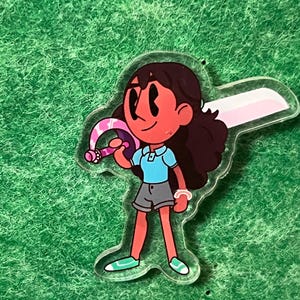 May include: A clear acrylic charm featuring a cartoon character with brown hair, wearing a blue shirt, gray shorts, and holding a pink and white sword. The charm is on a green textured background.
