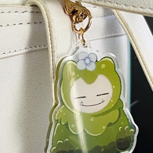 May include: A clear acrylic keyring featuring a green, monster-like character wearing a small white flower on its head. The character has a smiling expression with visible teeth and is attached to a gold-toned metal lobster clasp hook.