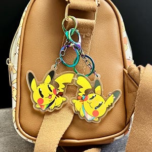 Puede incluir: Dos colgantes amarillos de Pikachu con detalles de arcoíris están sujetos a un bolso marrón con una correa beige. Los colgantes son de acrílico transparente y muestran al personaje en una pose dinámica. El bolso tiene una correa beige y un estampado blanco.