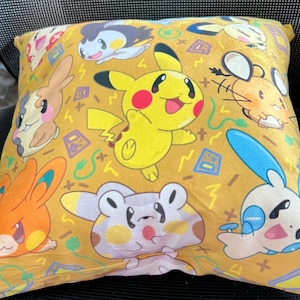 May include: Square cushion with a repeating pattern of cartoon characters on a mustard yellow background. The characters include Pikachu and other colorful creatures. The cushion is likely made of soft fabric.