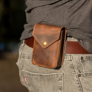 May include: A brown leather belt pouch with a brass button closure. The pouch is worn on a person's hip and is secured with a brown leather belt.