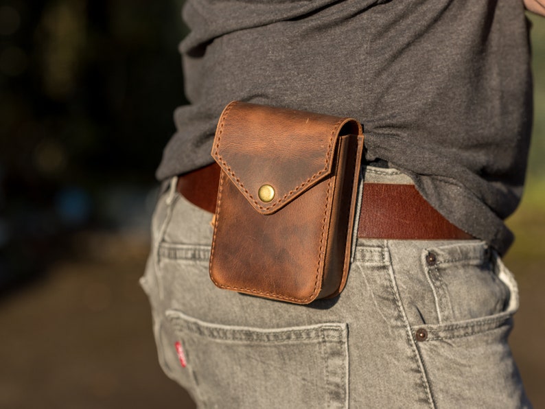 May include: A brown leather belt pouch with a brass button closure. The pouch is worn on a person's hip and is secured with a brown leather belt.