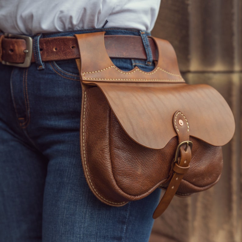 May include: A brown leather belt bag with a flap closure and a buckle. The bag is worn on a person's hip over a pair of blue jeans.