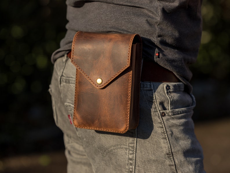 May include: A brown leather fanny pack with a flap closure and a brass button. The pack is worn on a person's waist.