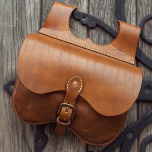 May include: A brown leather satchel bag with a buckle closure and a flap with a stitched design. The bag is hanging on a wooden surface.