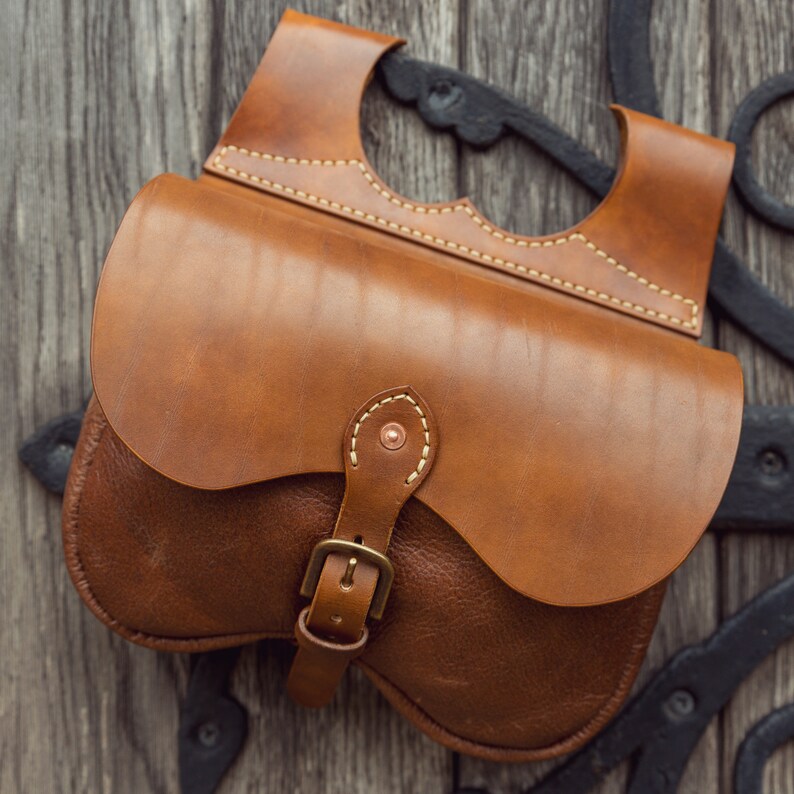 May include: A brown leather satchel bag with a buckle closure and a flap with a stitched design. The bag is hanging on a wooden surface.