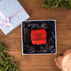May include: A red leather measuring tape in a white box with black tissue paper. The measuring tape has the text "LIFE'S TOO SHORT FOR BAD MEASUREMENTS". A light blue gift box with a purple ribbon is in the background.