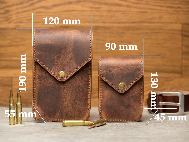 May include: Two brown leather phone cases with a flap closure and a brass button. The larger case measures 120 mm by 190 mm and the smaller case measures 90 mm by 130 mm. The cases are shown on a wooden surface with bullets and a belt.