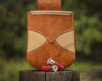 May include: A brown leather shotgun shell holder with a tan leather flap and stitching. The holder is sitting on a wooden surface with several shotgun shells in front of it. The text "Designed By - The House - Patterns UK" is visible on the holder.