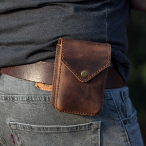 May include: A brown leather belt with a brown leather pouch attached to it. The pouch has a flap with a brass button closure. The belt is worn around the waist of a person wearing jeans.