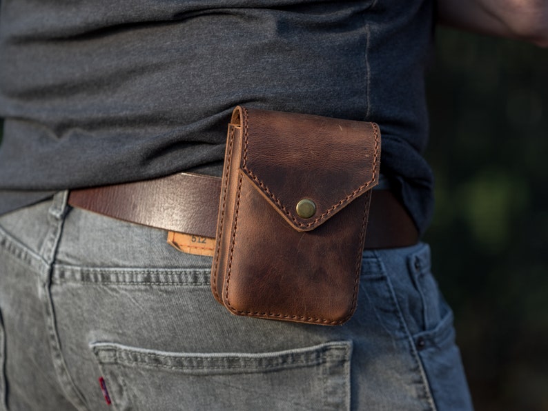 May include: A brown leather belt with a brown leather pouch attached to it. The pouch has a flap with a brass button closure. The belt is worn around the waist of a person wearing jeans.