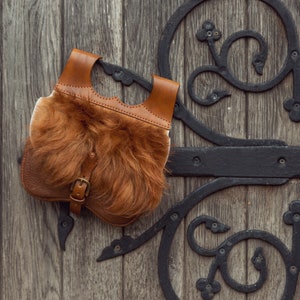 May include: A brown leather bag with a fur lining and a metal buckle. The bag is hanging on a wooden door with an ornate metal design.