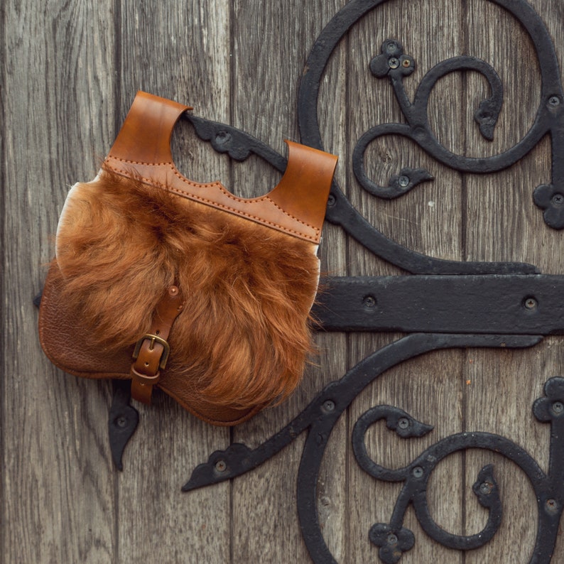 May include: A brown leather bag with a fur lining and a metal buckle. The bag is hanging on a wooden door with an ornate metal design.