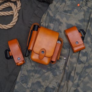 May include: A set of four brown leather pouches with metal clasps and loops. The pouches are arranged on a camouflage fabric background.