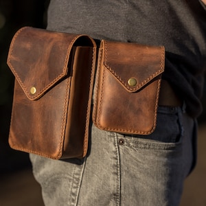 May include: Two brown leather pouches with brass snaps attached to a brown leather belt. The pouches are worn on a person's waist.