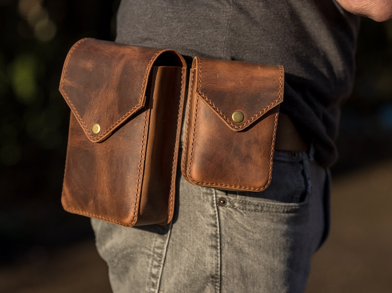 May include: Two brown leather pouches with brass snaps attached to a brown leather belt. The pouches are worn on a person's waist.