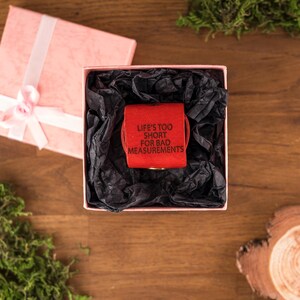 May include: A red measuring tape with the text "LIFE'S TOO SHORT FOR BAD MEASUREMENTS" inside a pink gift box with black tissue paper. The box is open, and a pink gift box with a white ribbon is next to it.