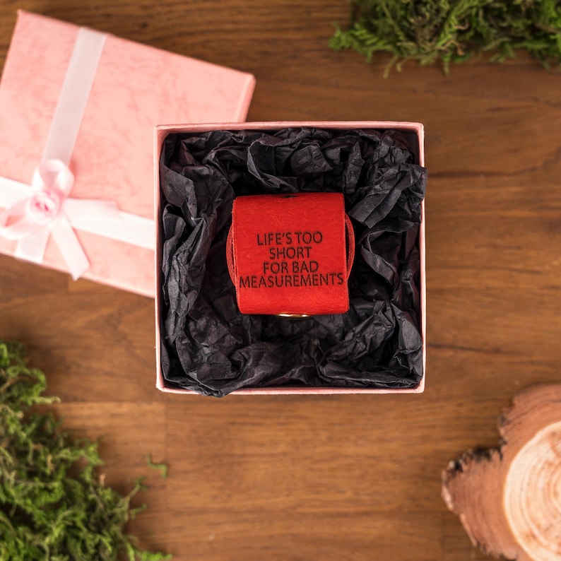 May include: A red measuring tape with the text "LIFE'S TOO SHORT FOR BAD MEASUREMENTS" inside a pink gift box with black tissue paper. The box is open, and a pink gift box with a white ribbon is next to it.