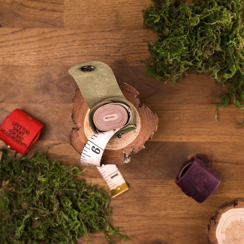 May include: A rolled measuring tape in a green leather case sits on a wooden slice, with the tape extending out. A red tape measure holder with the text "LIFE'S TOO SHORT FOR BAD MEASUREMENTS" is to the left. Moss and a purple leather holder are also visible.