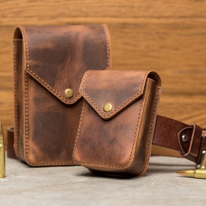 May include: Two brown leather pouches with a flap closure and a snap button. The smaller pouch is attached to a brown leather belt with a silver buckle. The pouches are on a light brown wooden surface.
