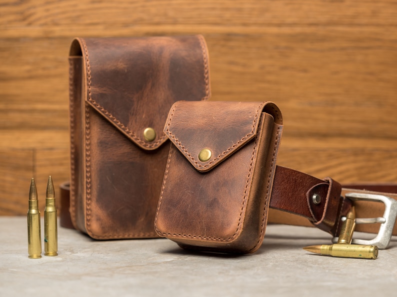 May include: Two brown leather pouches with a flap closure and a snap button. The smaller pouch is attached to a brown leather belt with a silver buckle. The pouches are on a light brown wooden surface.