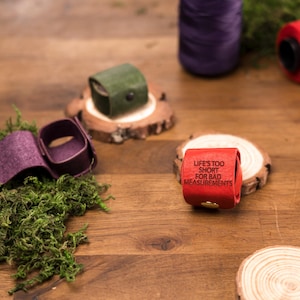 May include: A collection of leather measuring tape holders in various colors, including red, green, and purple. One red holder displays the text "LIFE'S TOO SHORT FOR BAD MEASUREMENTS". The holders are displayed on wooden slices.