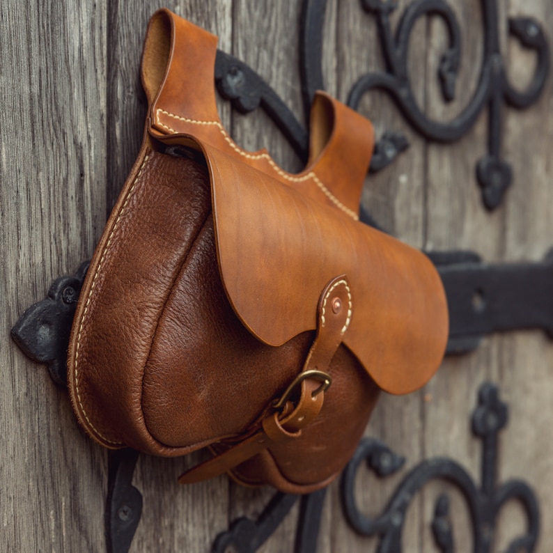 May include: A brown leather satchel with a flap and a buckle closure. The bag has a worn, vintage look and is hanging from a metal hook.