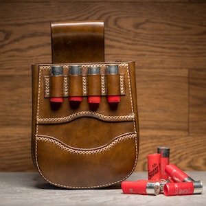 May include: A brown leather shell pouch featuring a belt loop and four vertical slots holding red shotgun shells. The pouch is constructed with visible light-colored stitching along the edges and decorative seams. Several red shotgun shells are scattered nearby.