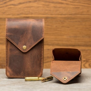 May include: Two brown leather pouches with a flap closure. The pouch on the left is closed, and the pouch on the right is open, revealing a brown leather interior. Three gold-colored bullets are in front of the pouches.