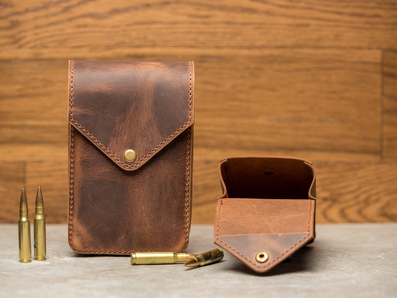 May include: Two brown leather pouches with a flap closure. The pouch on the left is closed, and the pouch on the right is open, revealing a brown leather interior. Three gold-colored bullets are in front of the pouches.