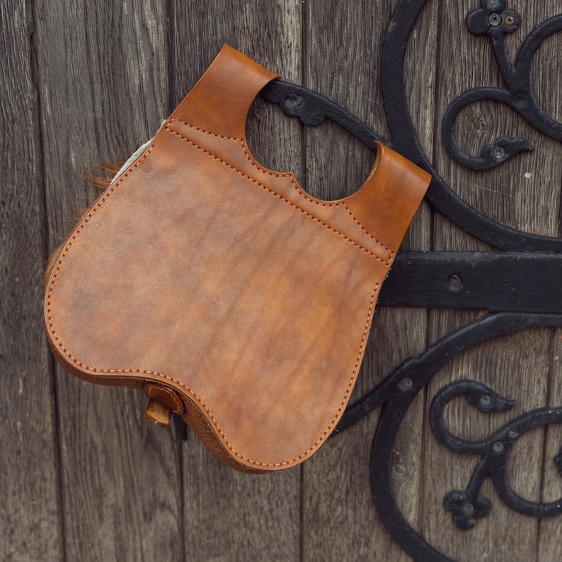 May include: A brown leather bag with a flap and a buckle closure. The bag is hanging on a wooden door with black metal accents.
