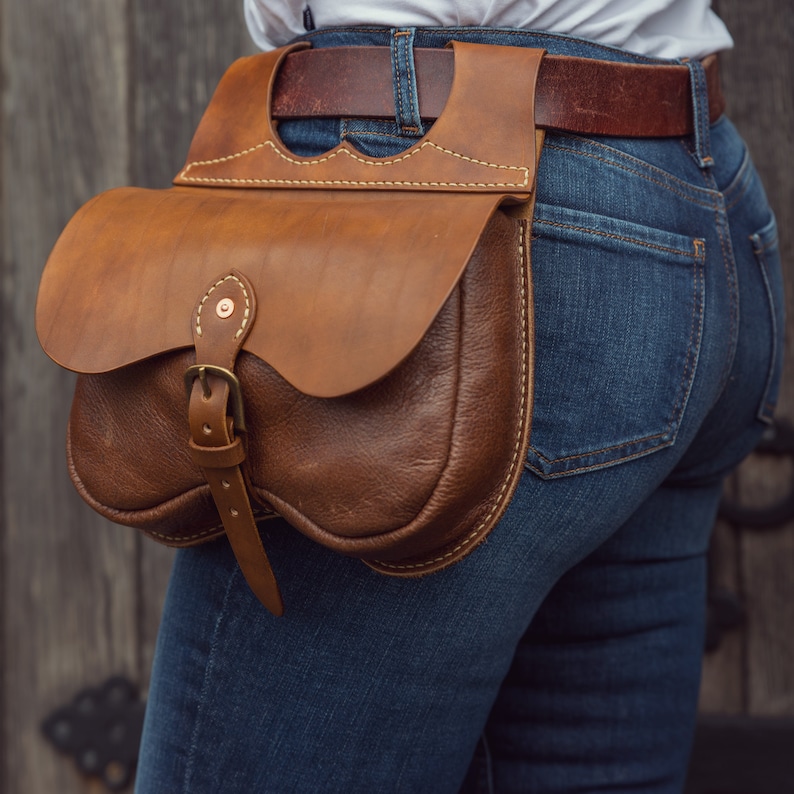 May include: A brown leather belt bag with a buckle and a strap. The bag is worn on a person's hip over a pair of blue jeans.