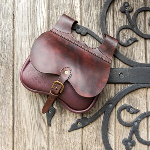 May include: A brown leather satchel with a flap closure and a brass buckle. The bag has a distressed look and is hanging on a wooden door.