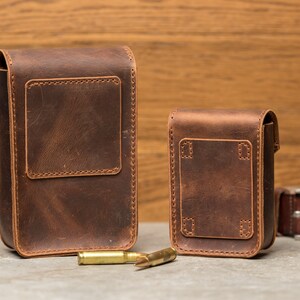 May include: Two brown leather pouches with stitched edges and flaps. The larger pouch is on the left and the smaller pouch is on the right. Both pouches have a rectangular shape and are made of a distressed leather. There are three gold-colored bullets in the foreground.