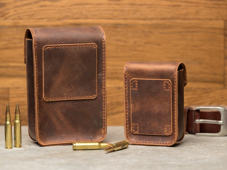 May include: Two brown leather pouches with stitched edges and flaps. The larger pouch is on the left and the smaller pouch is on the right. Both pouches have a rectangular shape and are made of a distressed leather. There are three gold-colored bullets in the foreground.