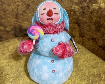 May include: A whimsical snowman figurine with a light blue body, a red scarf, and matching mittens. The snowman has a red nose and is holding a colorful lollipop. The figurine has a blue hood and is decorated with pink gems.