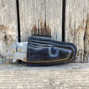 May include: A black leather knife sheath with a silver folding knife inside. The sheath has a belt loop and is made of high-quality leather.