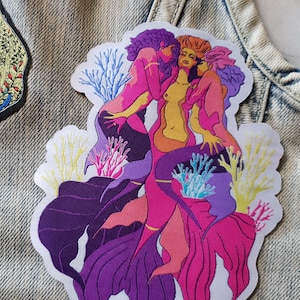Könnte beinhalten: Gestickter Aufnäher mit drei Meerjungfrauen mit lila, rosa und gelben Schwänzen, die sich ineinander verschlungen haben und von Korallen umgeben sind. Die Meerjungfrauen sind mit langen, fließenden Haaren dargestellt und umarmen sich.