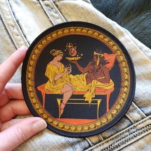 Könnte beinhalten: Ein runder schwarzer Patch mit goldener Umrandung, der zwei Figuren in einer Szene aus der griechischen Mythologie darstellt. Die Figuren sind eine Frau in einem gelben Gewand und ein Mann in einem braunen Gewand. Die Frau hält einen Teller mit einem roten Herzen und Sternen darauf. Der Hintergrund ist schwarz mit goldener Umrandung.