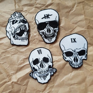 May include: Four white embroidered skull patches with black details. The skulls have different designs, including a rose, sunglasses, a pipe, and Roman numerals.