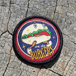 以下が含まれることがあります： 黒の縁取りと赤と白の縁取りが付いた丸いパッチ。中央には笑顔の顔をした漫画のハンバーガーと、黄色い文字で「we do BURGERS」と書かれている。