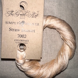 May include: A skein of pale peach-colored embroidery floss, labeled "Straw Bonnet 7002" by The Gentle Art. The floss is wound into a circular shape, with a cardboard tag attached. The tag has a hole for hanging.