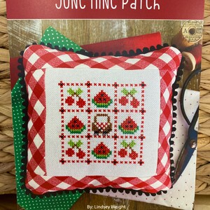 May include: A red and white gingham fabric pillow with a black trim. The pillow has a white cross-stitch design of watermelons, cherries, and a basket. The text "JUNE nine Patch" is at the top of the image. The text "By: Lindsey Weight" is below the pillow. The text "PRIMROSE cottage Stitches" is at the bottom of the image.