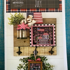 May include: A July calendar crate featuring a cross-stitch design of a house with fireworks and the text "Freedom Fireworks Made in the USA". The crate also includes a smaller cross-stitch design of a wreath with the text "Land that I Love".
